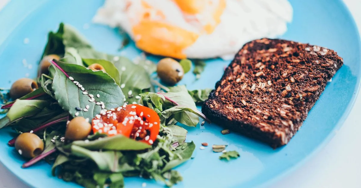 Un plato de desayuno nutritivo que presenta huevos, pan de centeno, ensalada y aceitunas en un vibrante plato azul.