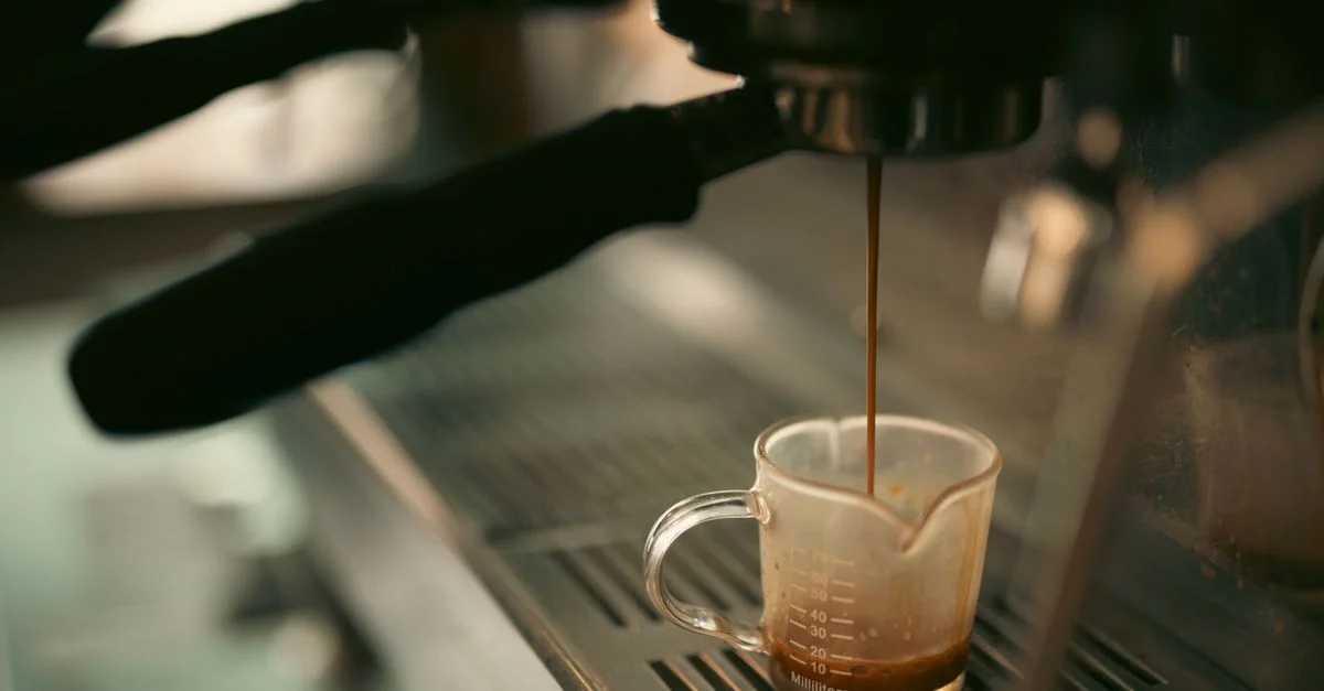 Café expreso vertido en una taza medidora desde una máquina de café, tonos cálidos.