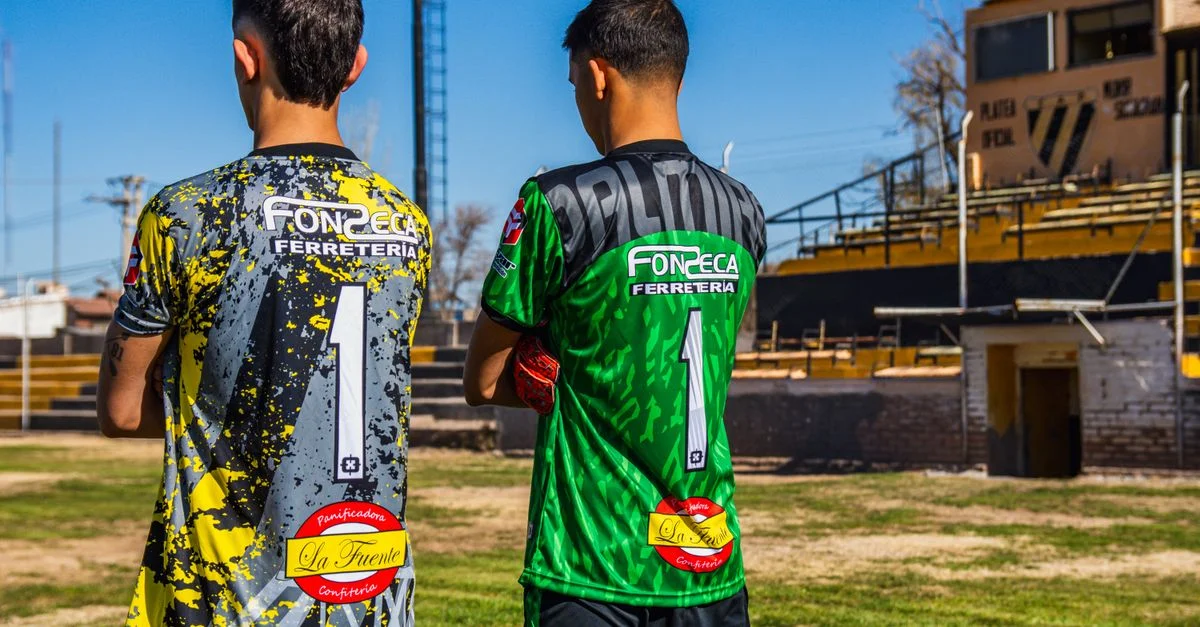 Dos jóvenes futbolistas con camisetas coloridas, listos para un partido en un campo urbano.