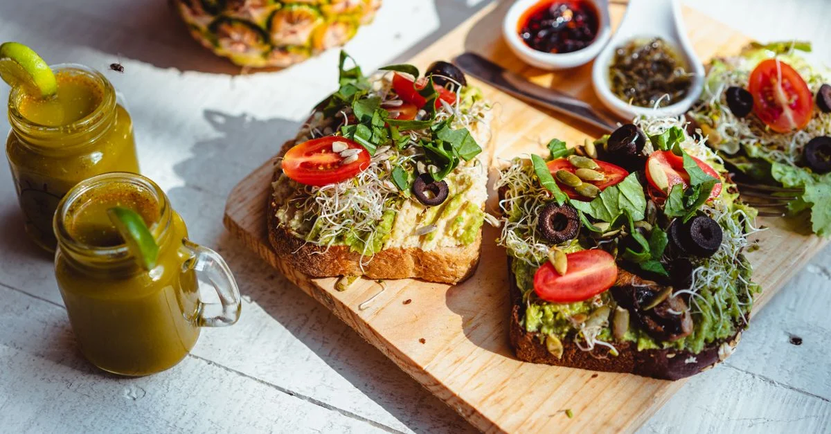 Primer plano de tostada de aguacate fresca con coberturas, servida con jugos en una tabla de madera.