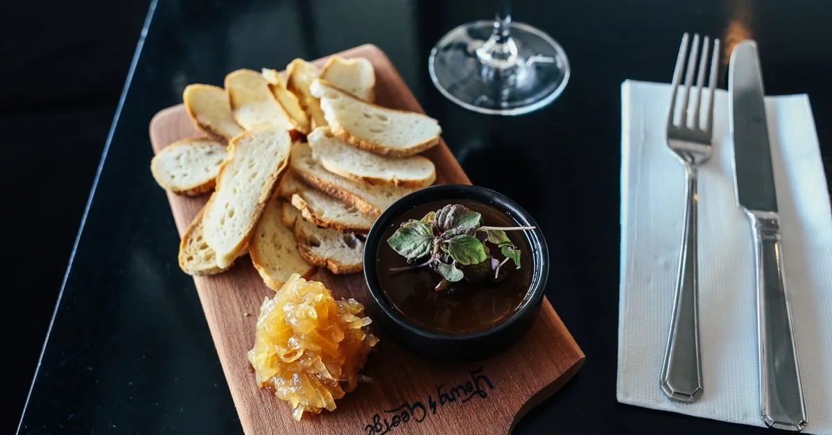 Una sofisticada bandeja de quesos y panes con chutney servida elegantemente al lado de una copa de vino tinto en una mesa negra.