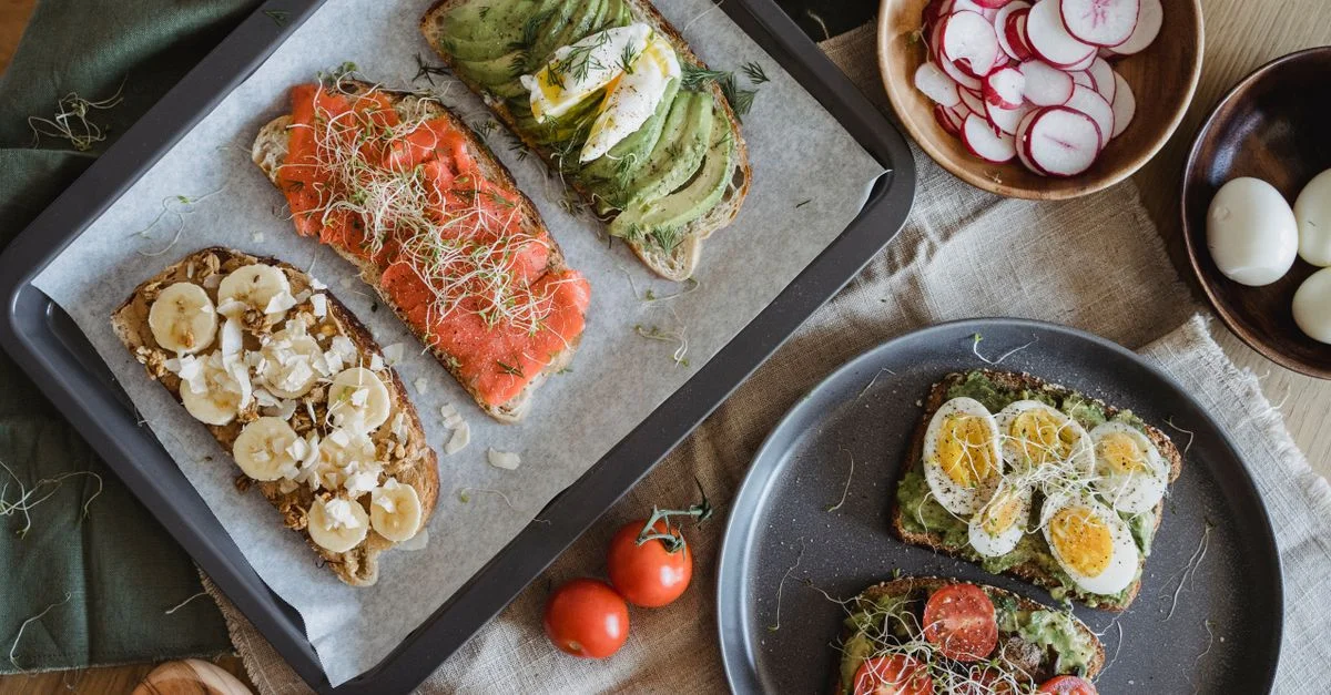 Una variedad de tostadas saladas y dulces con aguacate, plátano, huevos y coberturas frescas sobre una mesa de madera.