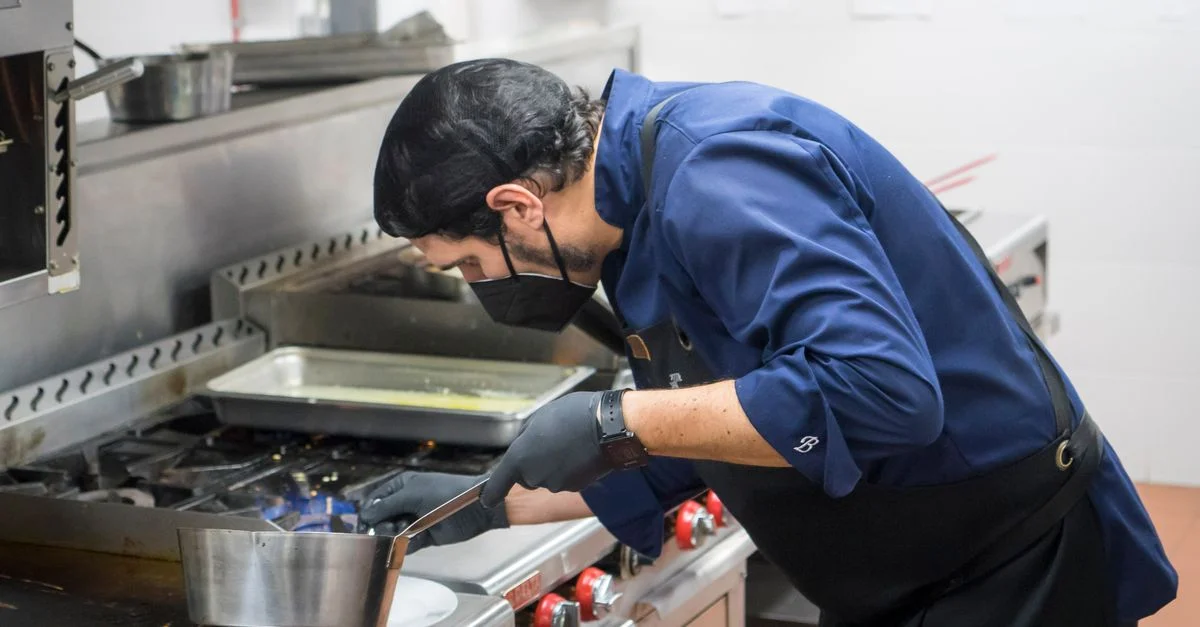 Chef con chaqueta azul cocinando un plato con precisión en la estufa de una cocina de restaurante.