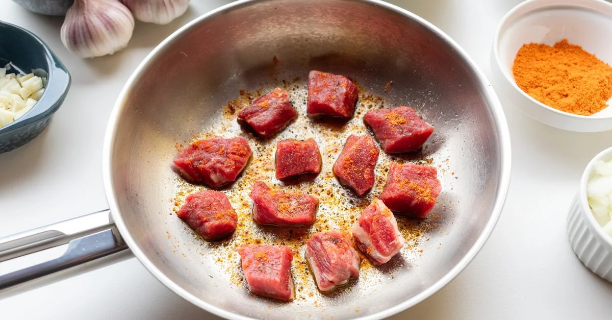 Trozos de carne cruda sazonada y sellada en una sartén rodeada de ingredientes frescos.