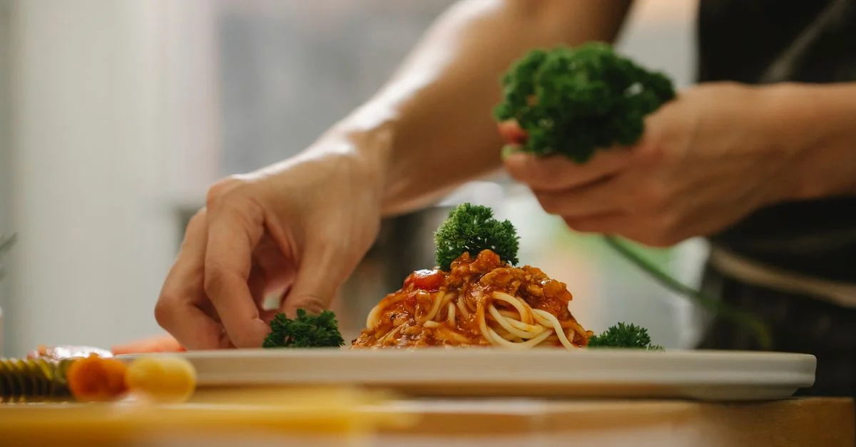 Primer plano de un chef adornando espaguetis con perejil en un entorno de cocina.