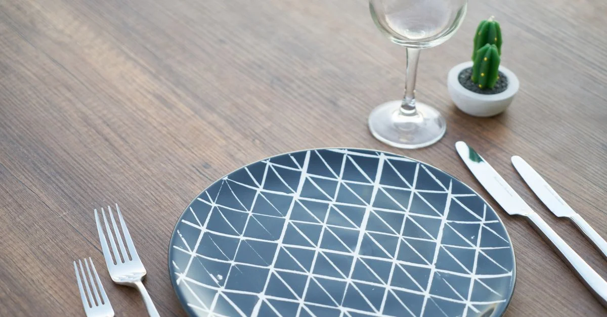 Configuración de mesa elegante con plato geométrico, cubiertos y cactus en maceta.