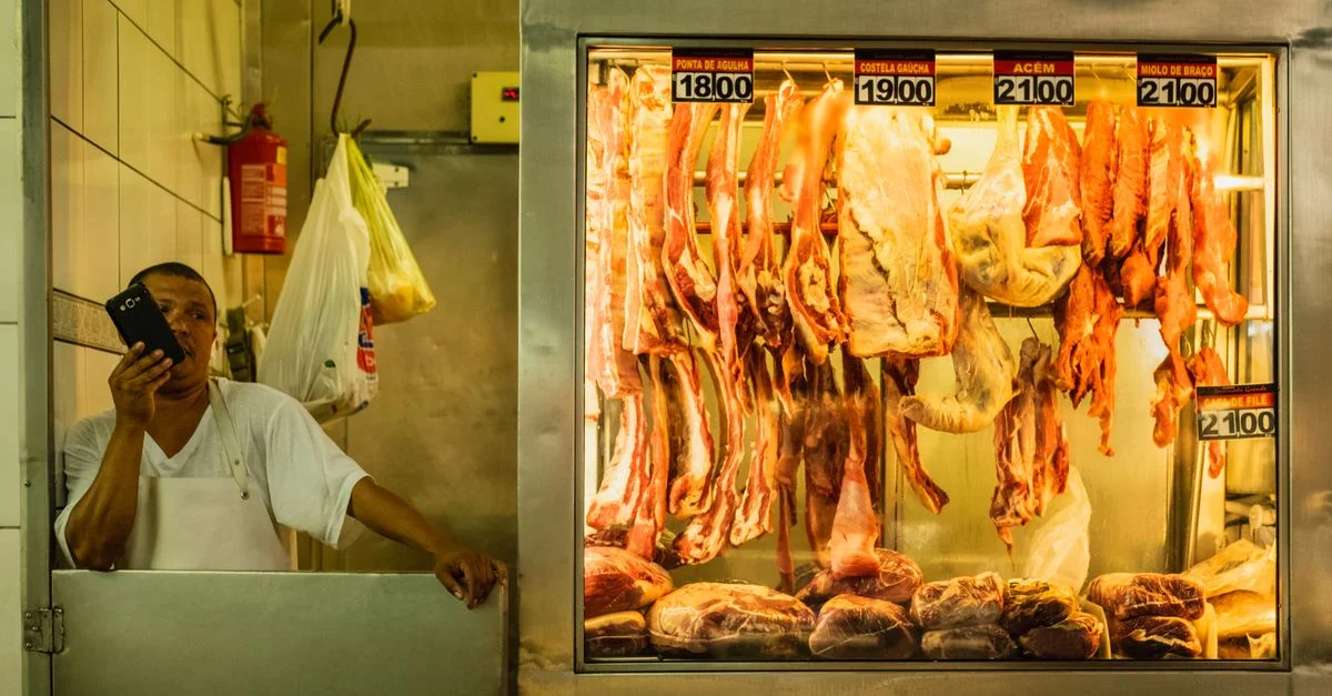Hombre en una carnicería usando el teléfono junto a un exhibidor de carnes colgadas con etiquetas de precio.