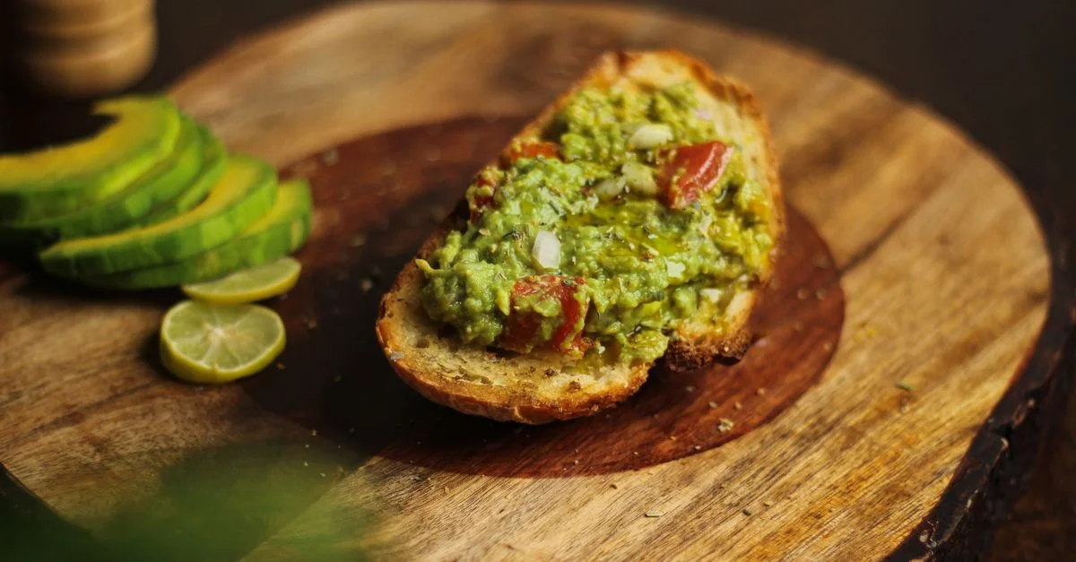 Primer plano de tostada de aguacate en una tabla de madera con aguacates en rodajas, destacando un bocado gourmet.