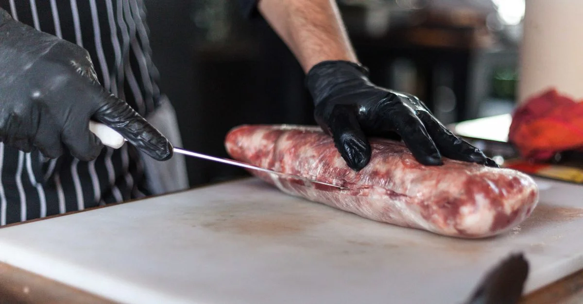 Un chef portant des gants découpe de la viande crue sur une planche à découper dans une cuisine.