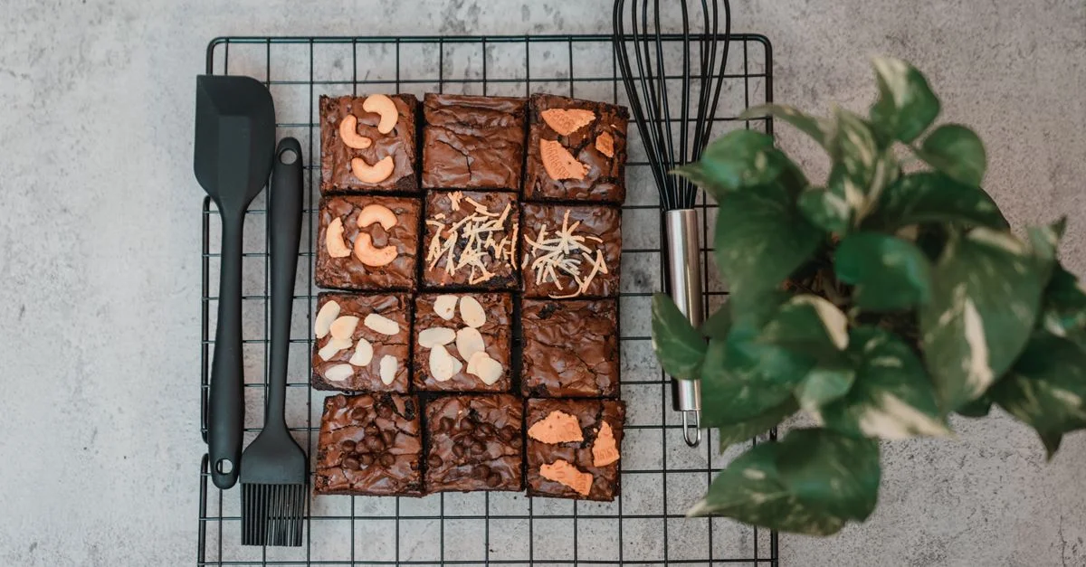 Una vista cenital de brownies caseros en una rejilla de enfriamiento con una planta y utensilios de cocina.