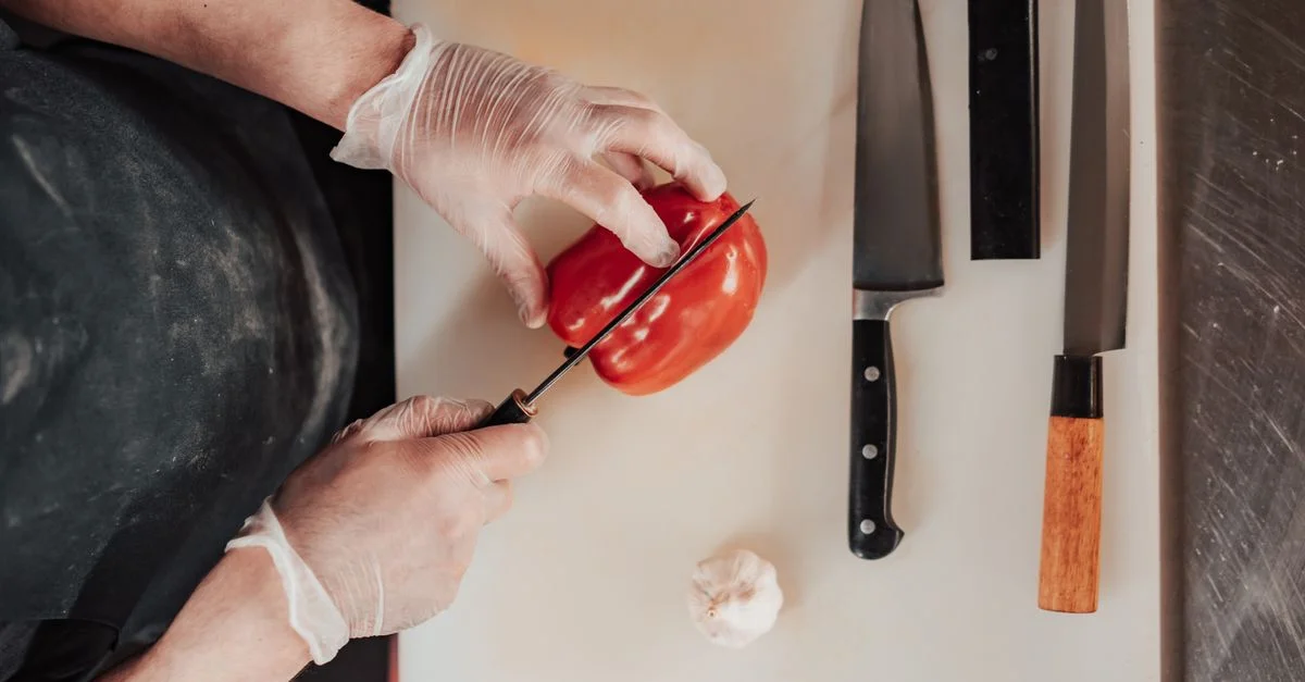 Vista superior de un chef cortando un pimiento rojo en una tabla de cortar con cuchillos, mostrando habilidades culinarias.