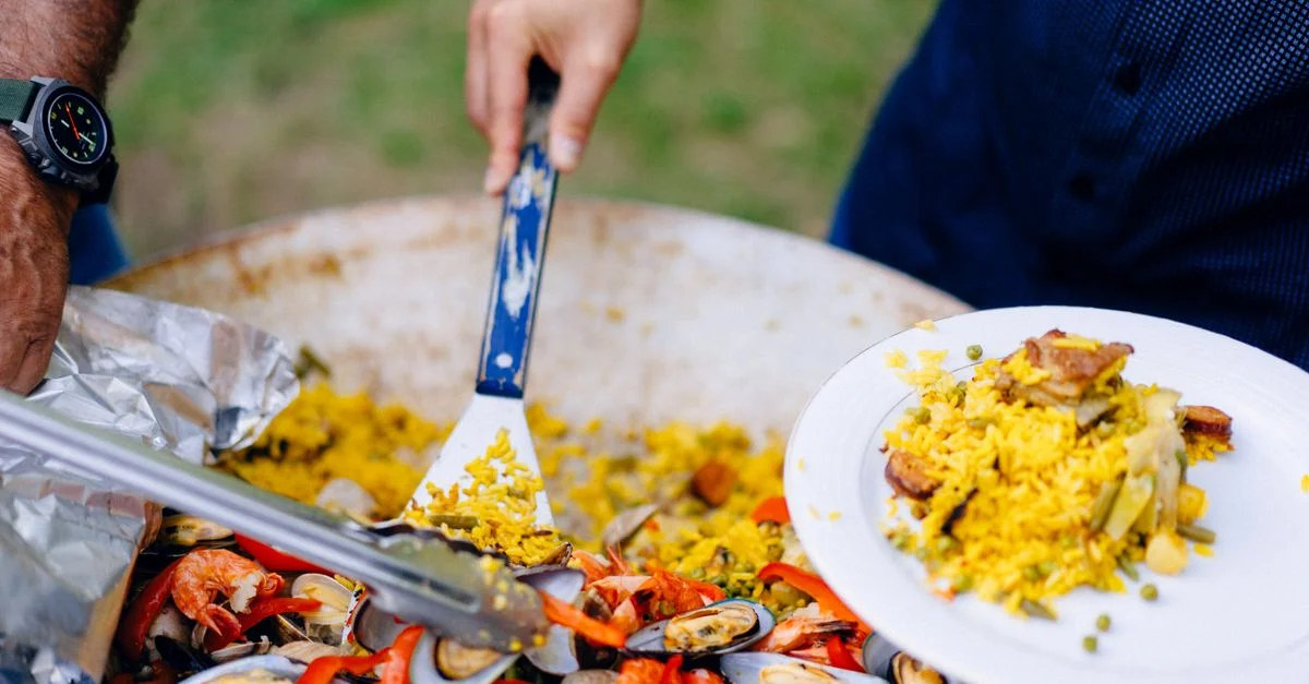 Un primer plano de manos sirviendo paella española con gambas y mejillones, destacando colores vibrantes e ingredientes deliciosos.