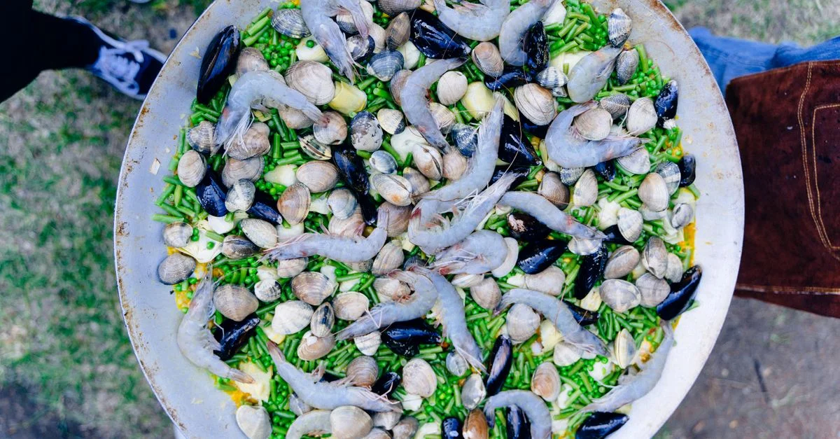 Primer plano de una paella de mariscos cruda con almejas, mejillones y camarones en una gran sartén al aire libre. Perfecto para viajes culinarios.