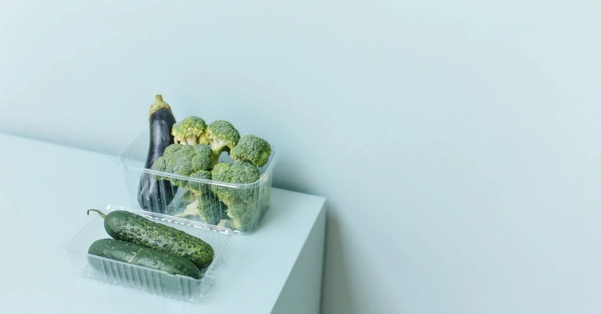 Surtido de verduras frescas como brócoli, berenjena y pepino en embalaje de plástico sobre una superficie blanca.