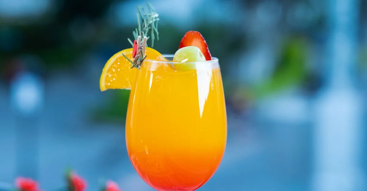 Un vibrante cóctel veraniego adornado con naranja, fresa y hierbas aromáticas, servido al aire libre.